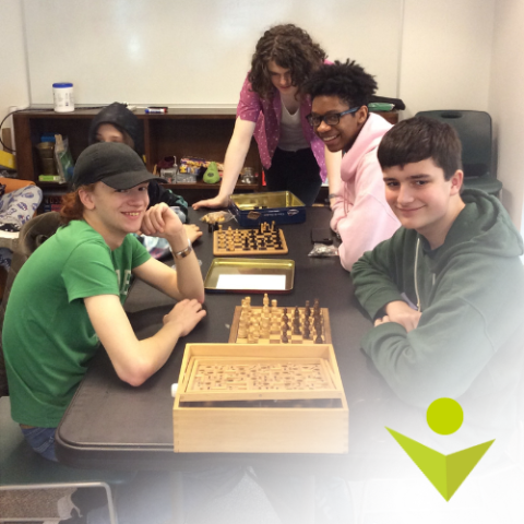 Several teens look smiling toward the camera as they play chess around a table at center