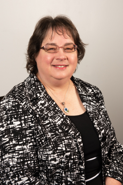 White woman with brown shoulder-length hair, glasses, and a black and white crosshatched blazer