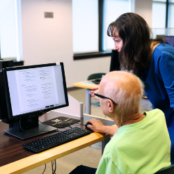 Person sitting at computer with another person leaning over to help.