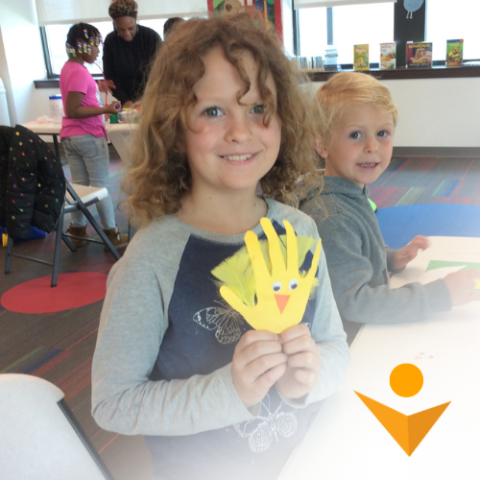 Boy with shoulder-length curly hair holds a yellow hand-shaped spring peep craft as other children work on their crafts in the background