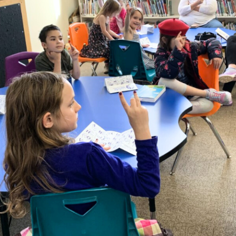 Children sitting at blue tables making sign language letters with hands.