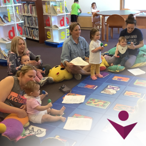 Infants and their caregivers sitting on the floor along a colorful rug with book shelves in the background