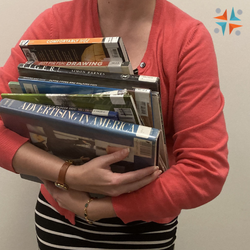 Woman holding a stack of books in a salmon color sweater with the WCFL logo in corner